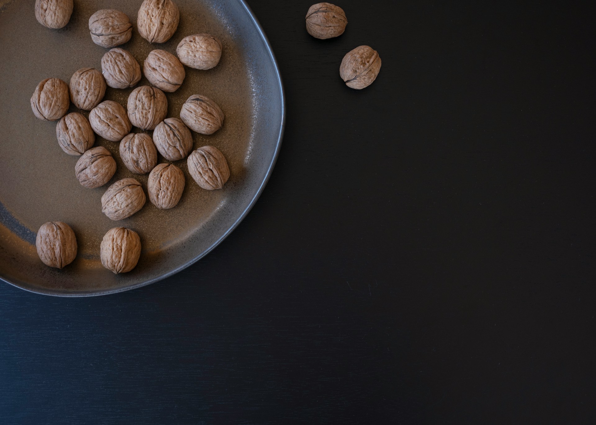 Whole walnuts in their shells on a glazed stoneware plate, shot from above against dark painted wood, with two nuts sitting apart on the bare surface