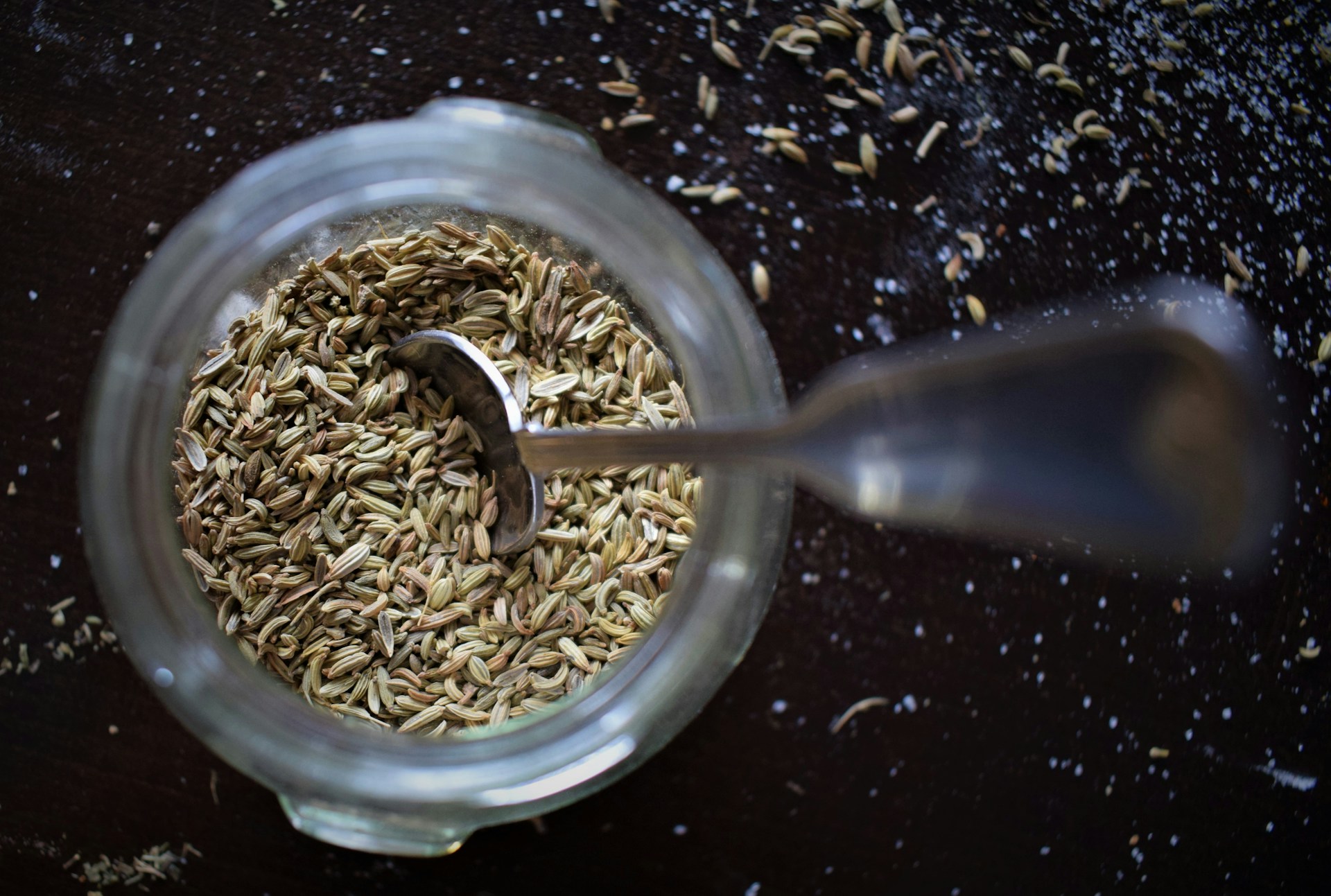 A glass jar of fennel seed shot from above with a metal spoon resting in it, seeds and salt scattered across the dark surface around it