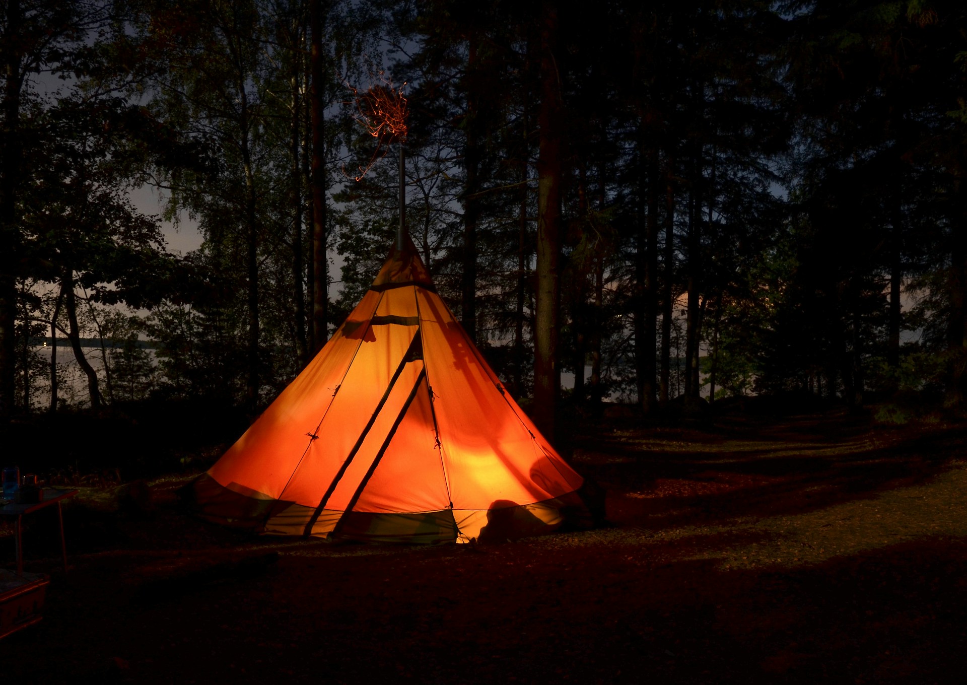 A lit tipi tent glowing orange in a pine forest at dusk, sparks rising from its stove pipe, a pale lake visible through the trunks behind