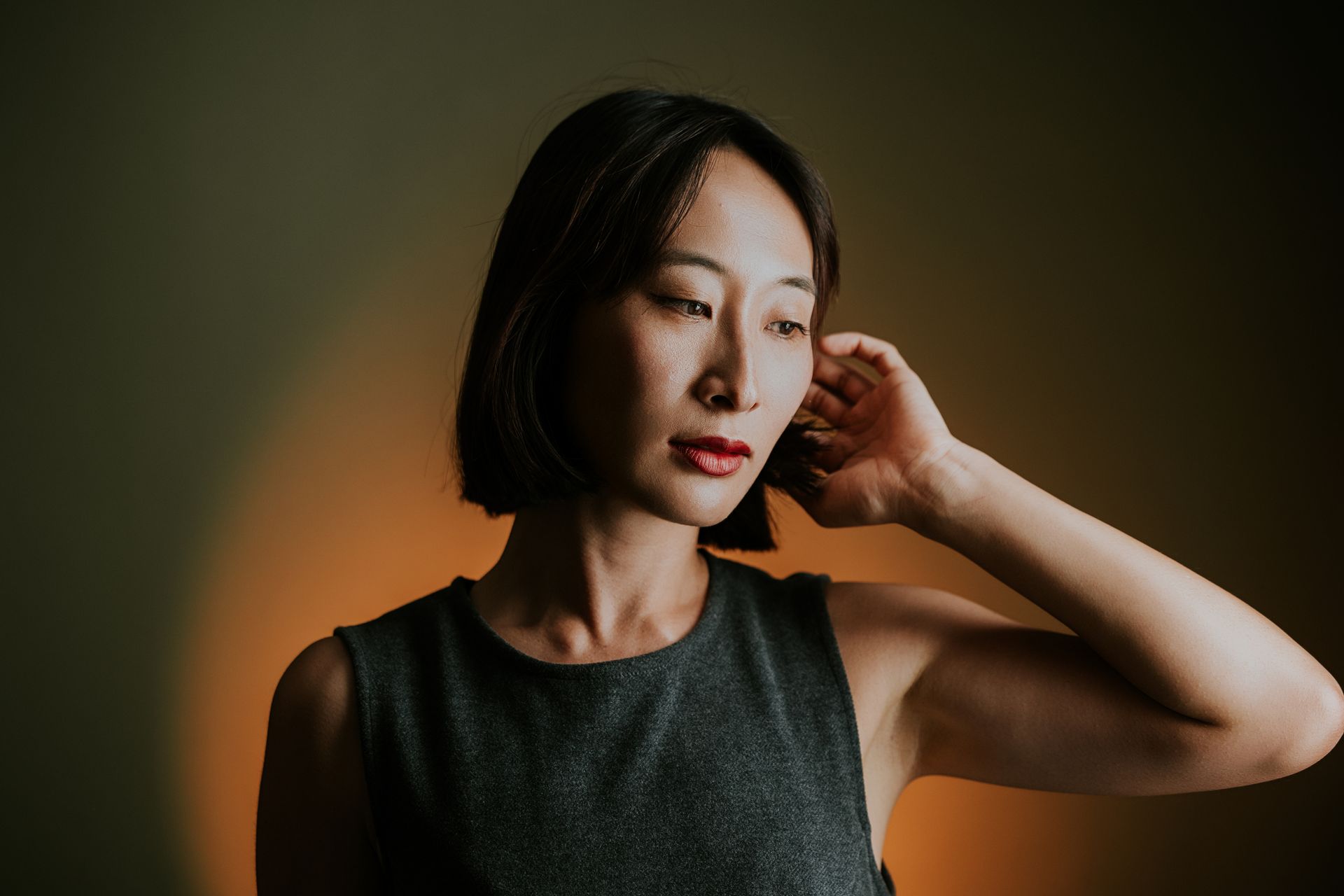 A woman with a short dark bob, one hand raised to her hair, standing in front of a warm amber wall in low light