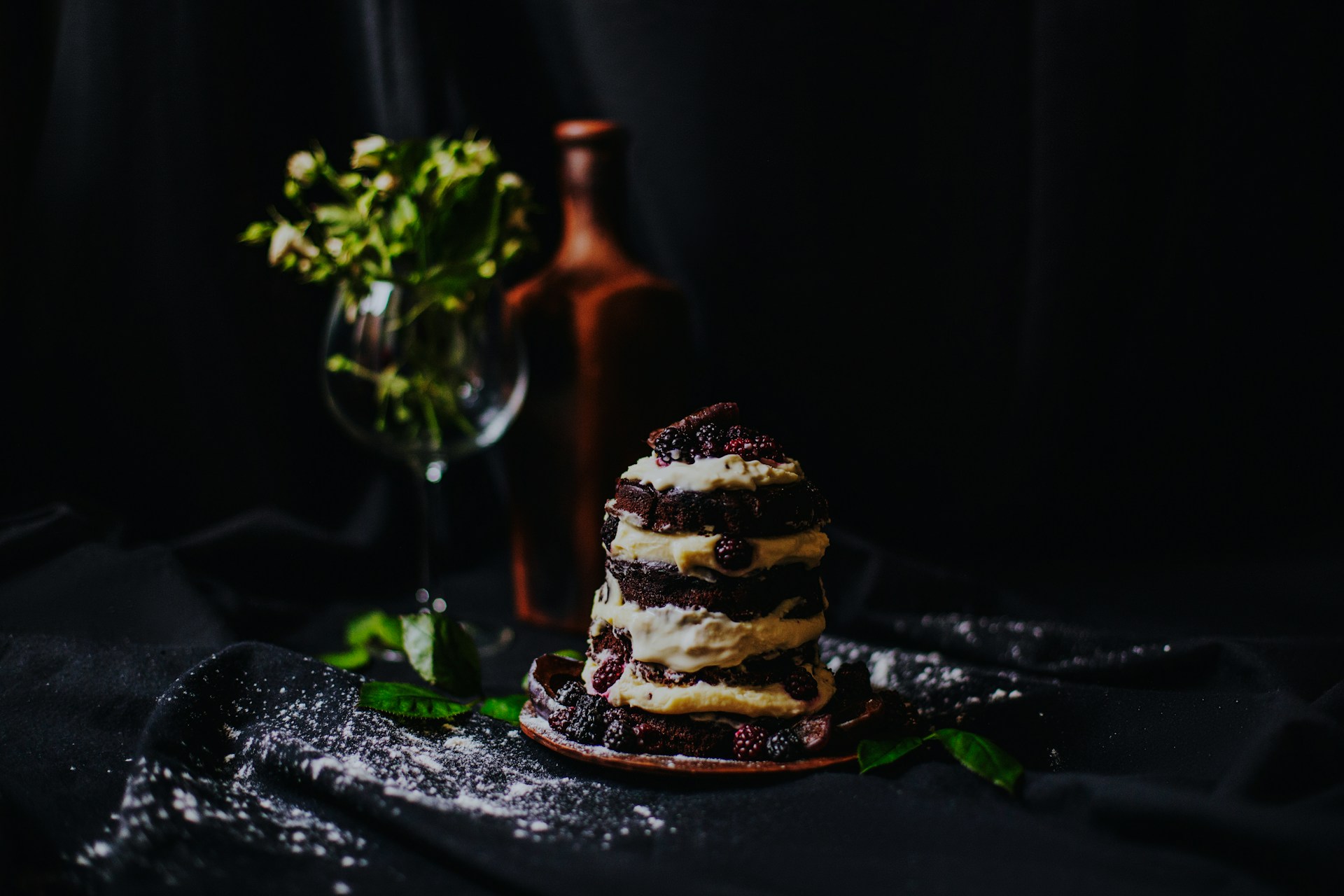 A small stacked layer cake of dark sponge, cream and blackberries on a clay plate, lit against a near-black backdrop with sugar dusted across the cloth