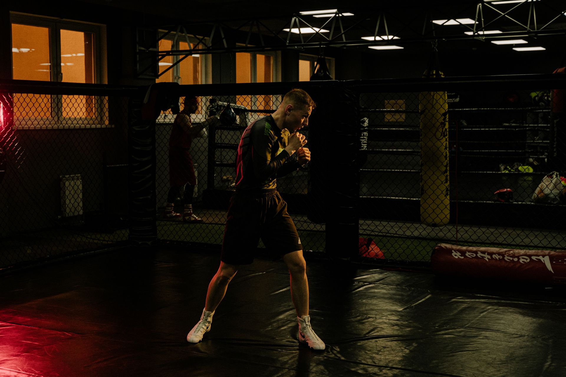 A fighter shadow-boxing alone inside a caged training floor, lit from one side, with heavy bags and lit windows in the dark behind