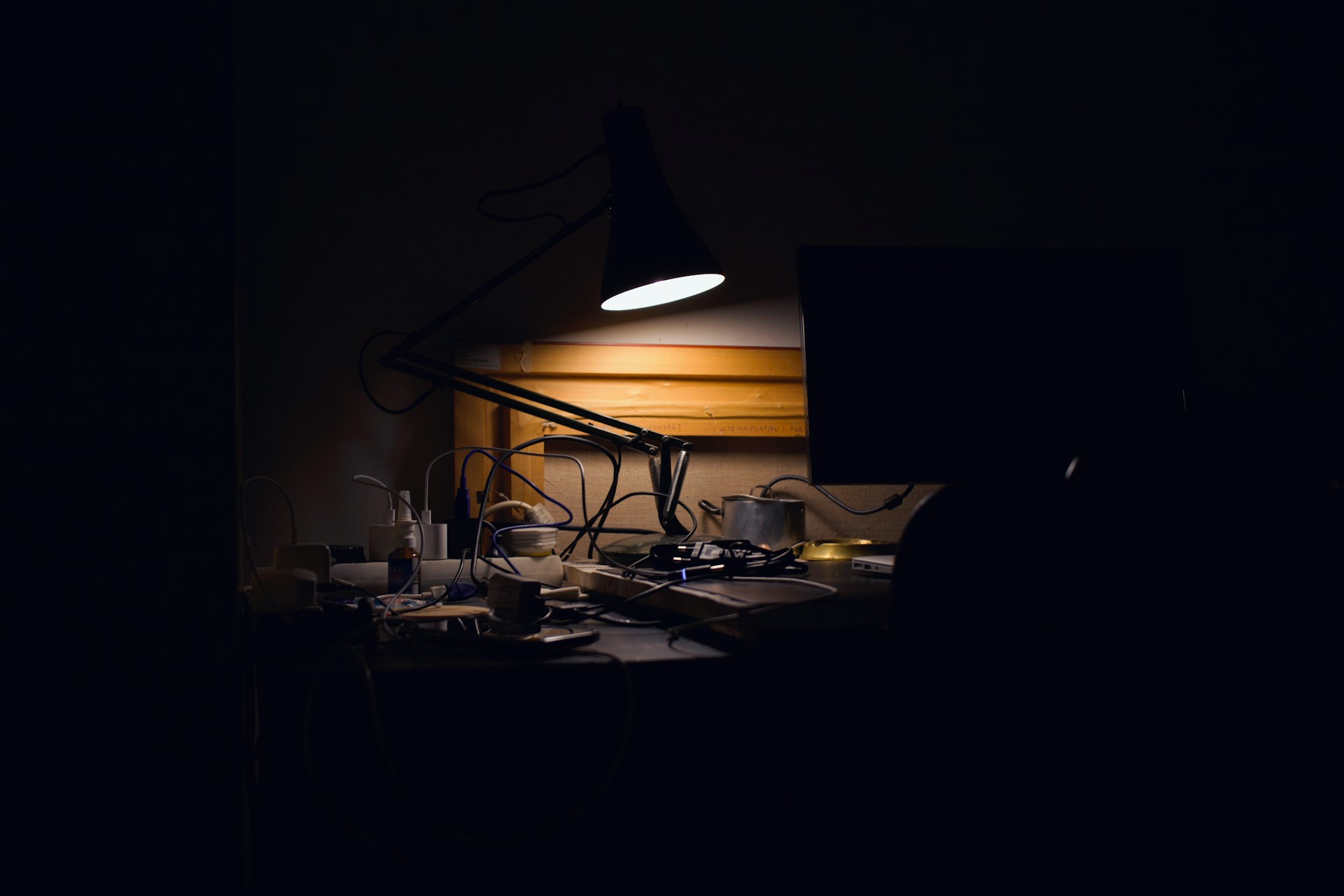 A cluttered desk late at night lit by one anglepoise lamp: tangled cables, a mug, small bottles and a dark monitor, with the rest of the room in blackness