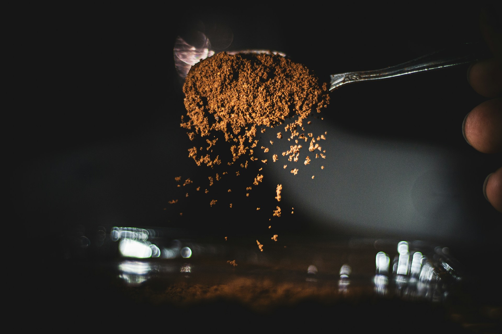 A spoonful of coarse brown powder tipped in mid-air, grains scattering down through a dark frame lit from one side